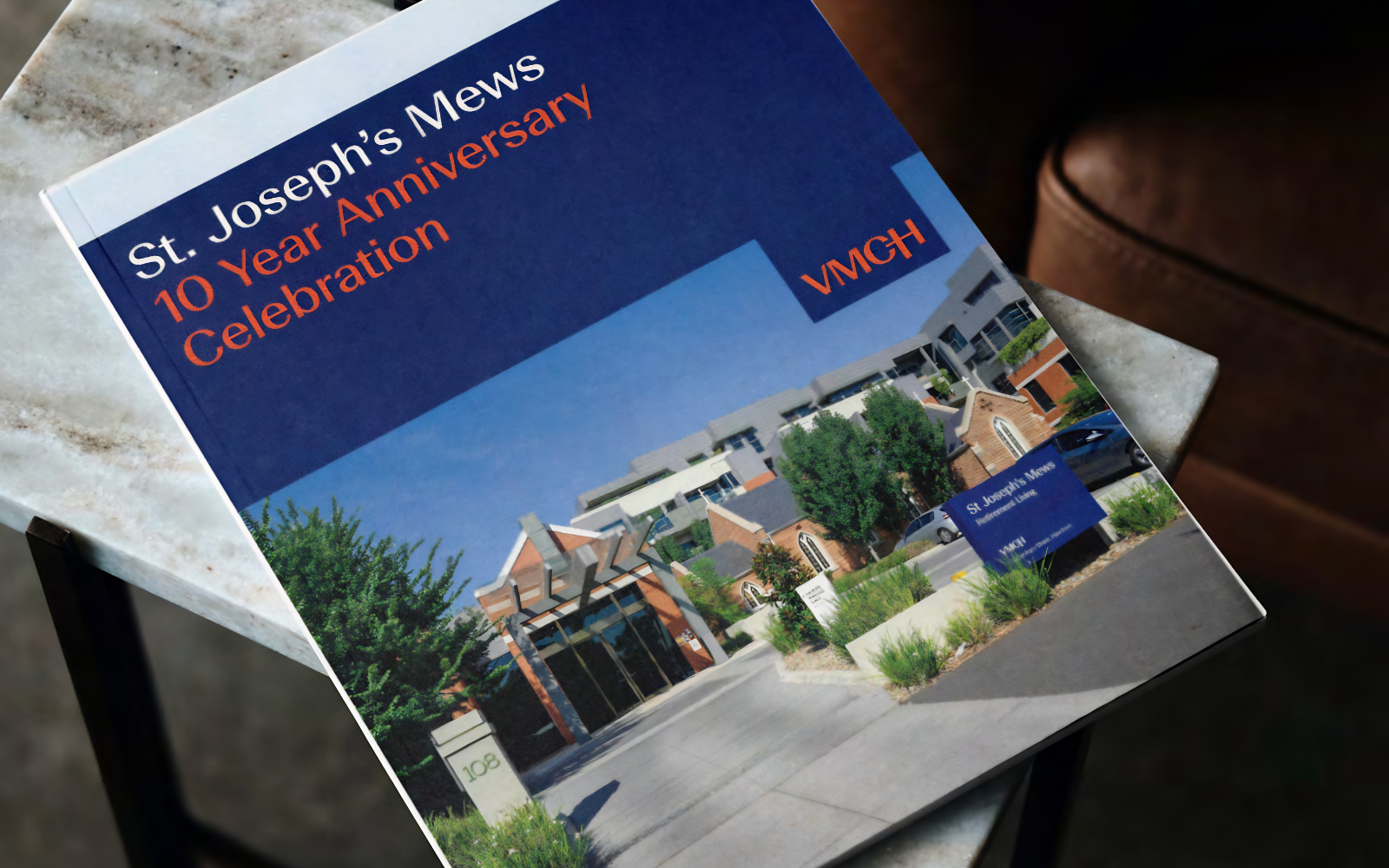 A brochure celebrating the 10 year anniversary of a retirement living development sits on top of a marble coffee table in front of a brown leather armchair